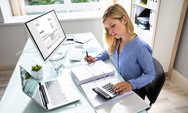 A woman using a calculator at a desk in front of a couple of computers