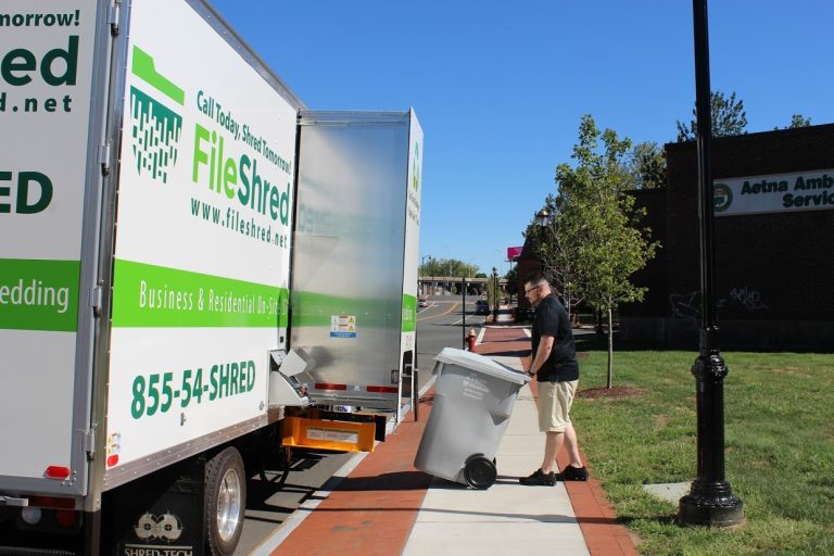 A white shredding truck with green and black text advertising "FileShred" and its services. The text includes the website "www.fileshred.net" and phone number "855-54-SHRED." The truck is parked on a gravel area near some buildings.