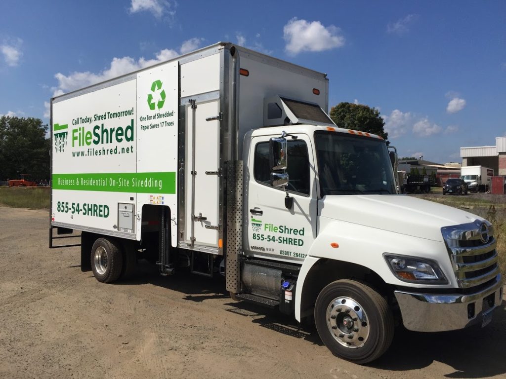 A white shredding truck with green and black text advertising "FileShred" and its services. The text includes the website "www.fileshred.net" and phone number "855-54-SHRED." The truck is parked on a gravel area near some buildings.