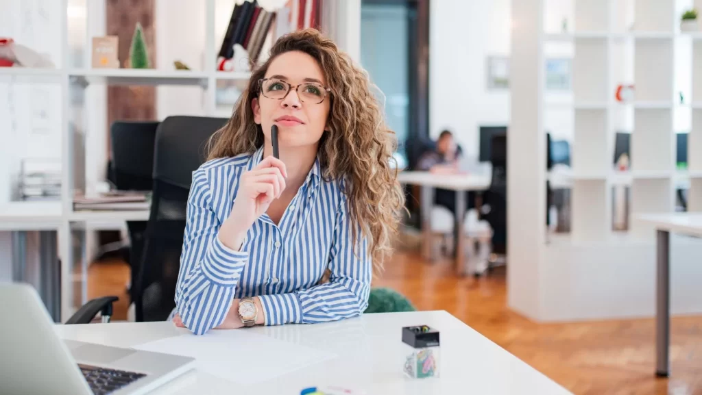 Woman in stripped shirt sitting at a desk with a pen to her chin in a thinking pose.