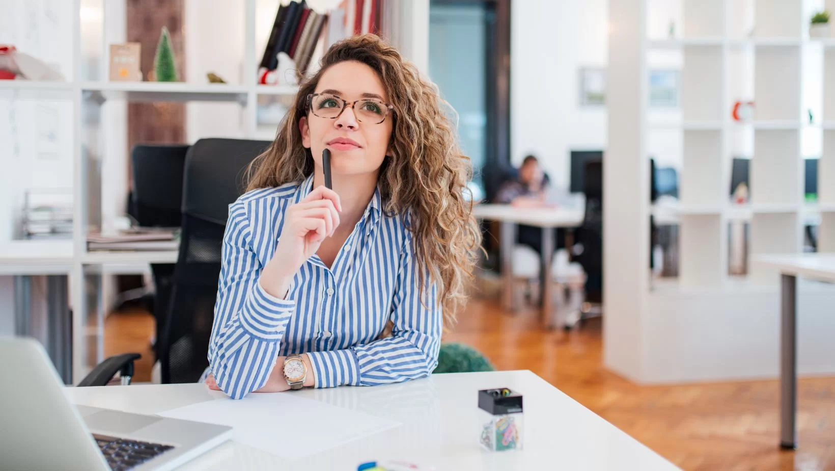 Woman in stripped shirt sitting at a desk with a pen to her chin in a thinking pose.