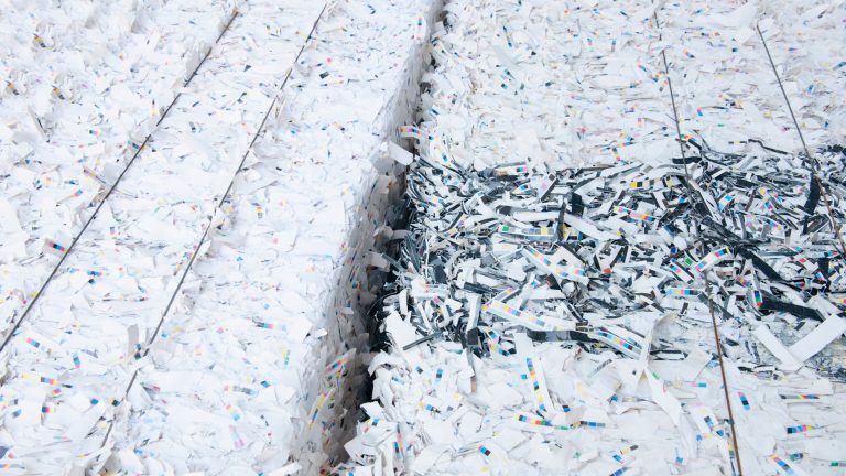 Large stacks of shredded white paper with colorful specks sit closely packed. One section has darker strips, creating a contrast in the otherwise pale scene.
